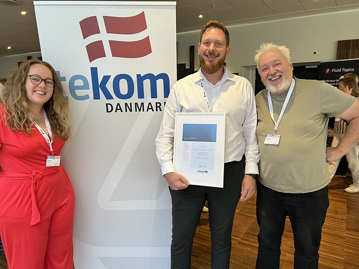 award winner Esben Keller in white shirt holding an award certificate standing next to award committee members Maiken Blok and Boo Engstrand standing next to the tekom Danmark roll-up banner